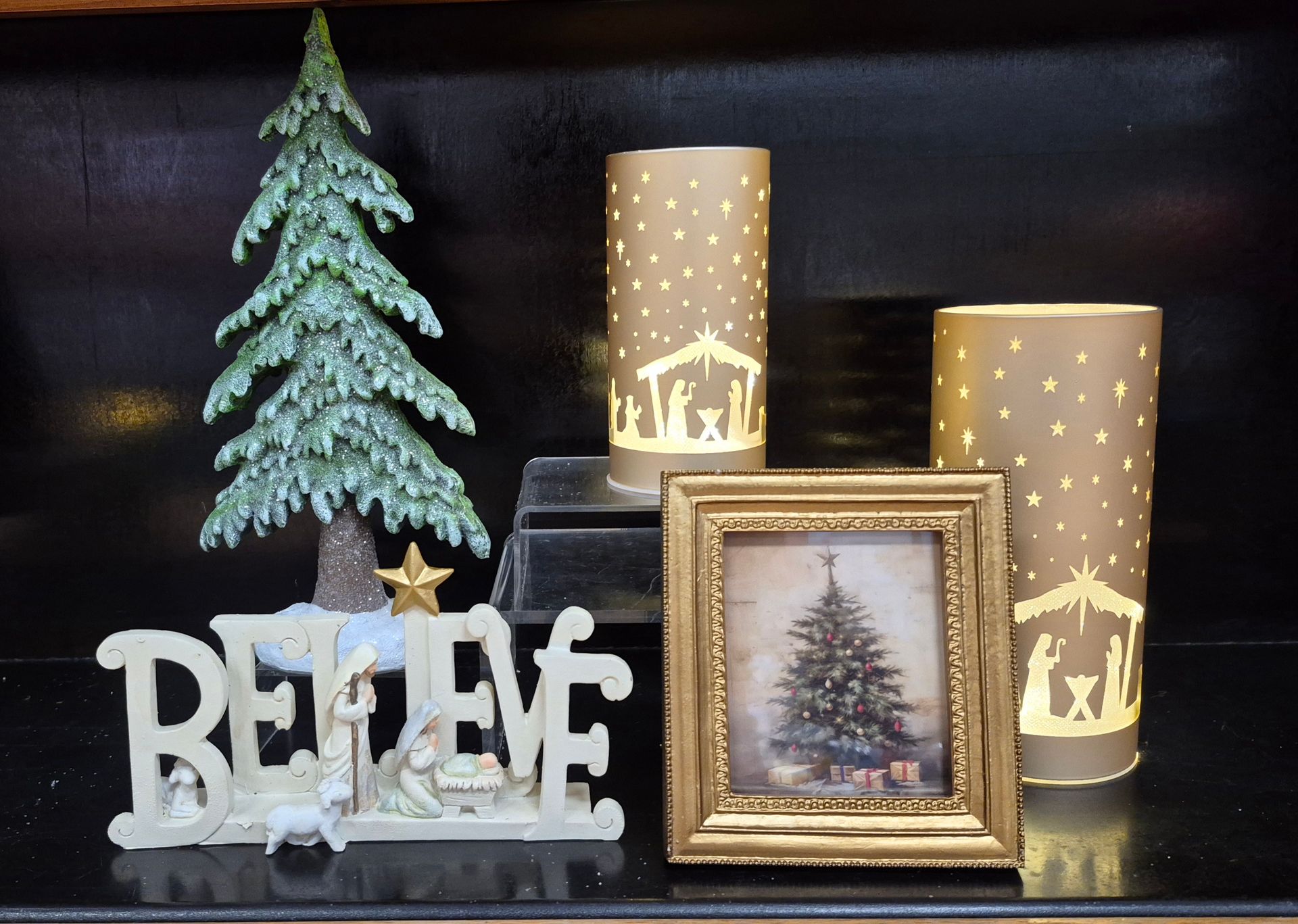Christmas shelf display: tree, BELIEVE nativity, framed tree art, two illuminated Bethlehem scene lanterns.