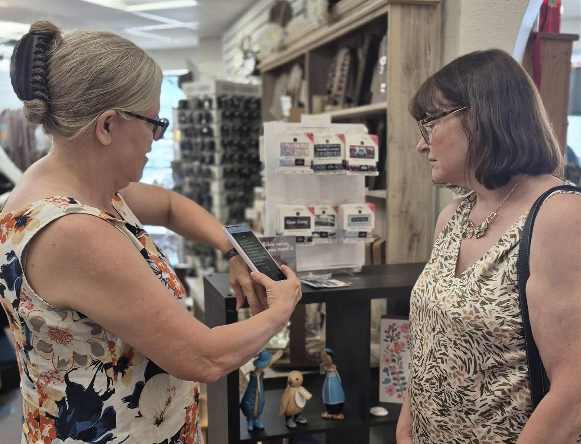 Woman taking photo of merchandise with phone, woman next to her looking at products. Store setting.