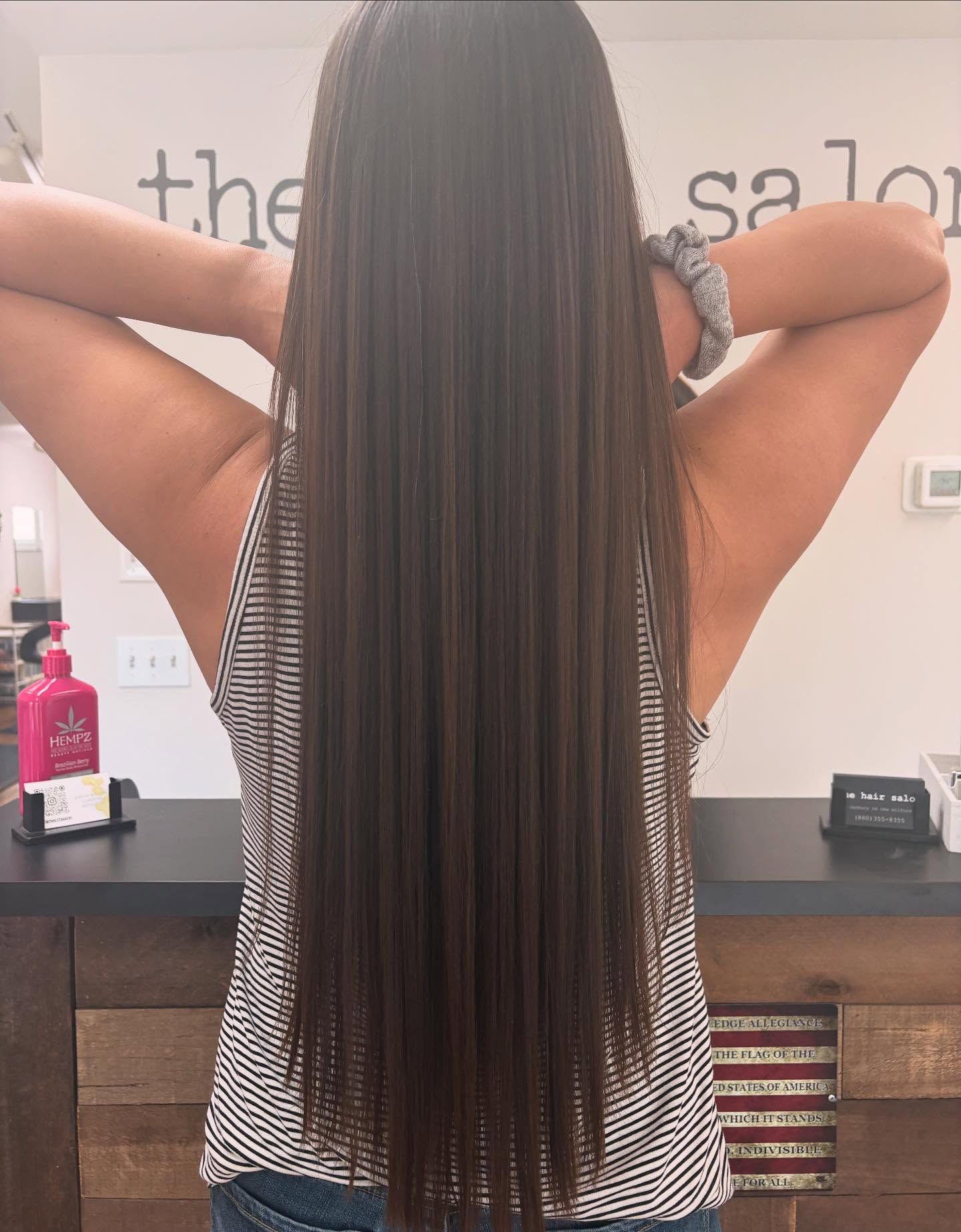 Person with long, straight brown hair in a salon, arms raised behind head.