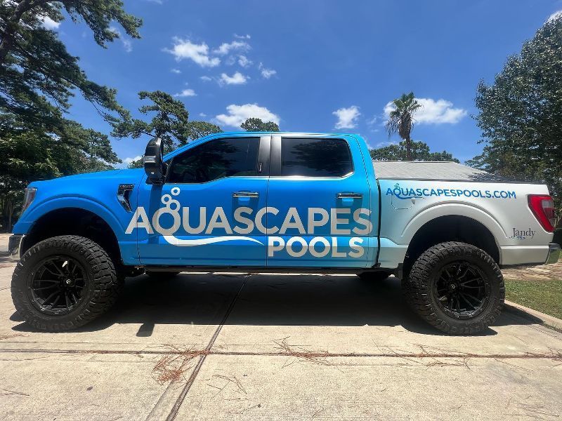 A blue truck with the words aquascapes pools on the side is parked in a driveway.