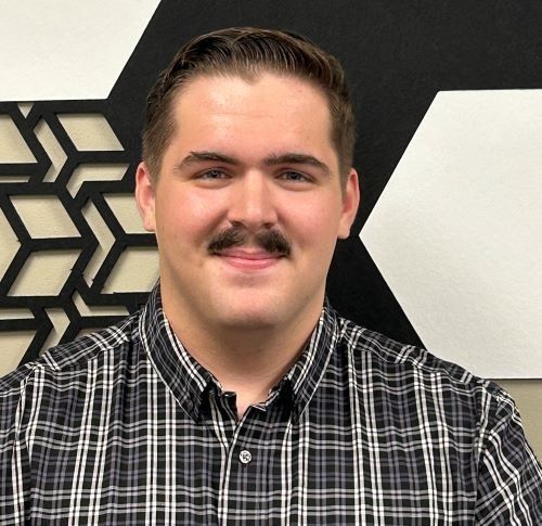 Man with mustache smiling, wearing a plaid shirt, in front of a geometric black and white wall.