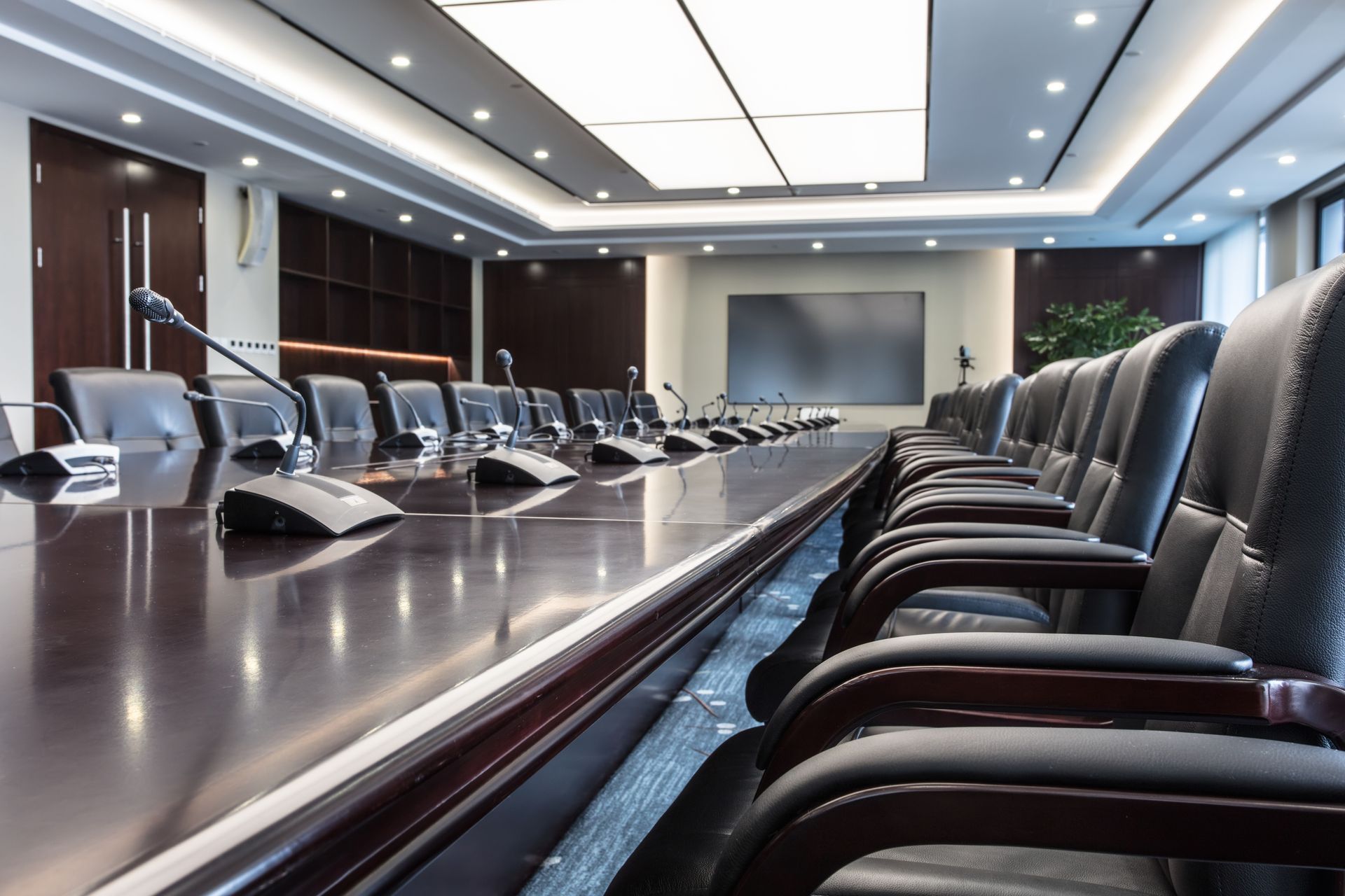 Conference room with long wooden table, chairs, microphones, and large screen.