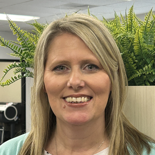 Woman with blonde hair smiling, standing in front of greenery.