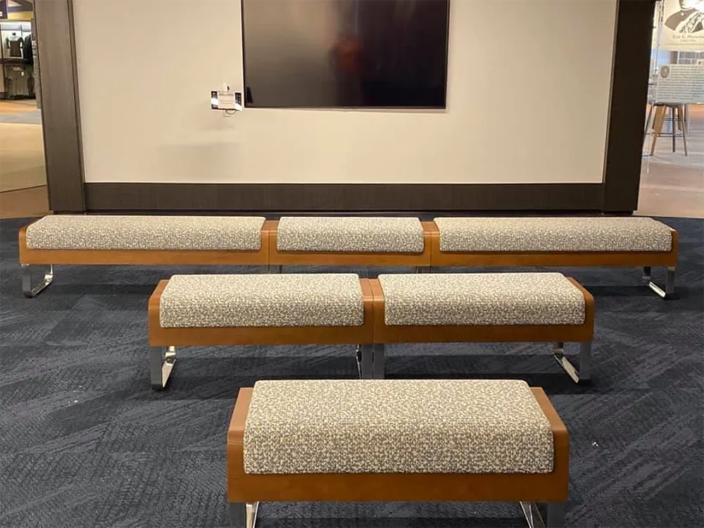 Five wood and speckled-fabric benches in front of a gray wall with a television and dark blue carpet.