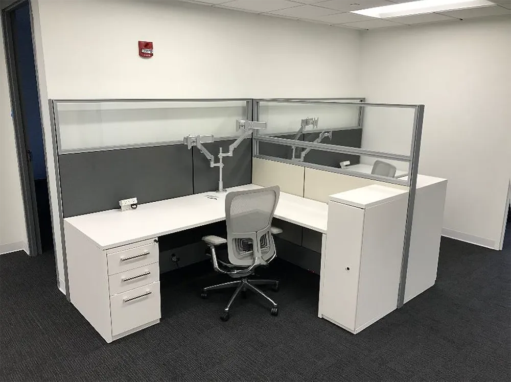 L-shaped office cubicle with white desk, gray walls, computer monitor arms, and a gray office chair.