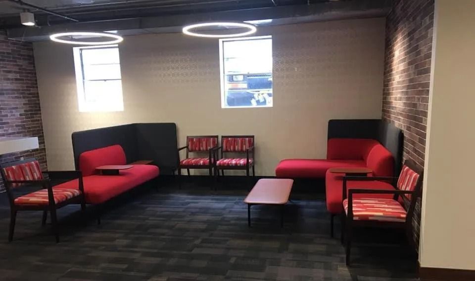 Lounge area with red couches and chairs, brick walls, and two windows. Gray carpet and circular lights overhead.
