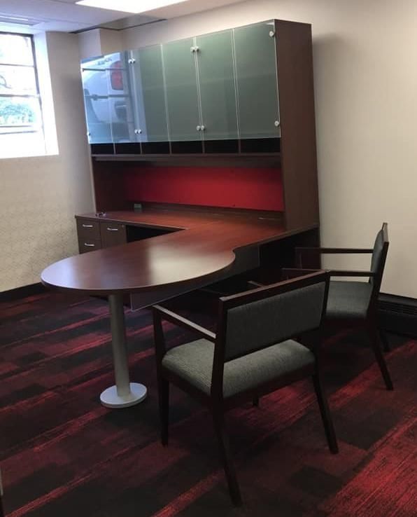 Office with a large dark wood desk, overhead storage, and two grey chairs. Red and black patterned carpet.