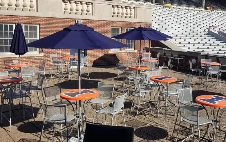 Outdoor seating area with tables, chairs, and umbrellas. Building and stadium seating in the background.