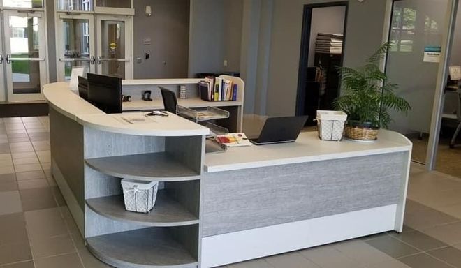Reception desk in a modern office lobby; gray and white curved counter with shelves, open doorway in background.