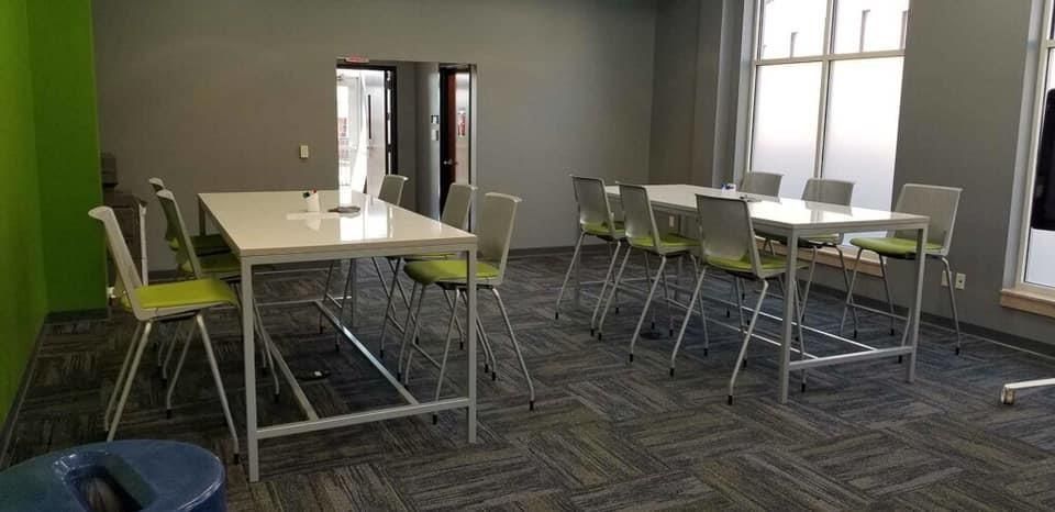 A room with two white tables, green and gray chairs, and windows.