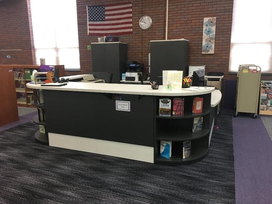 Library circulation desk with shelves of books, brick wall, and American flag.