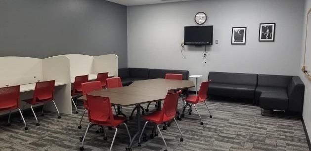 A room with desks, chairs, and couches. A television and wall art are mounted.