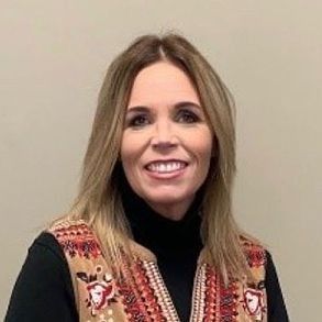 Woman with shoulder-length blonde hair, smiling, wearing a black turtleneck and patterned vest, indoors.