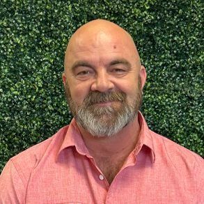Bald man with a grey beard and a pink shirt smiles in front of a green leafy backdrop.