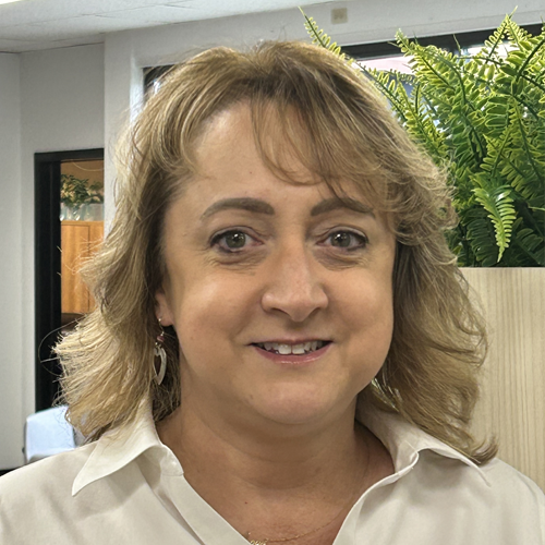 Woman with wavy blonde hair smiling, wearing a cream blouse, in an office setting.