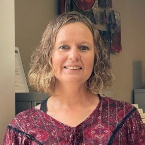Woman with short curly hair, smiling, wearing a burgundy patterned top, in an office setting.