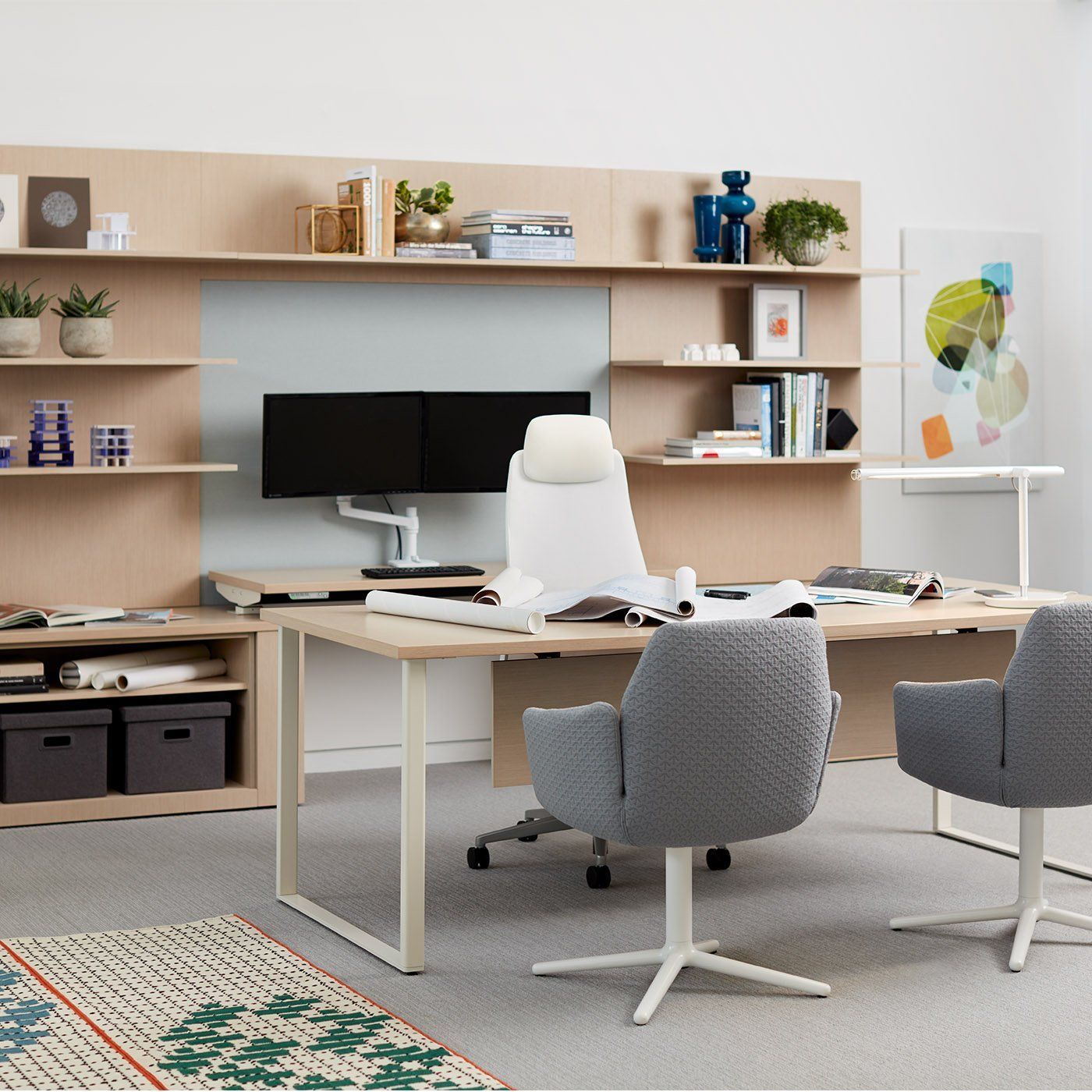 Modern office with light wood desk, two monitors, white chair, and two gray armchairs. Shelves hold decor.