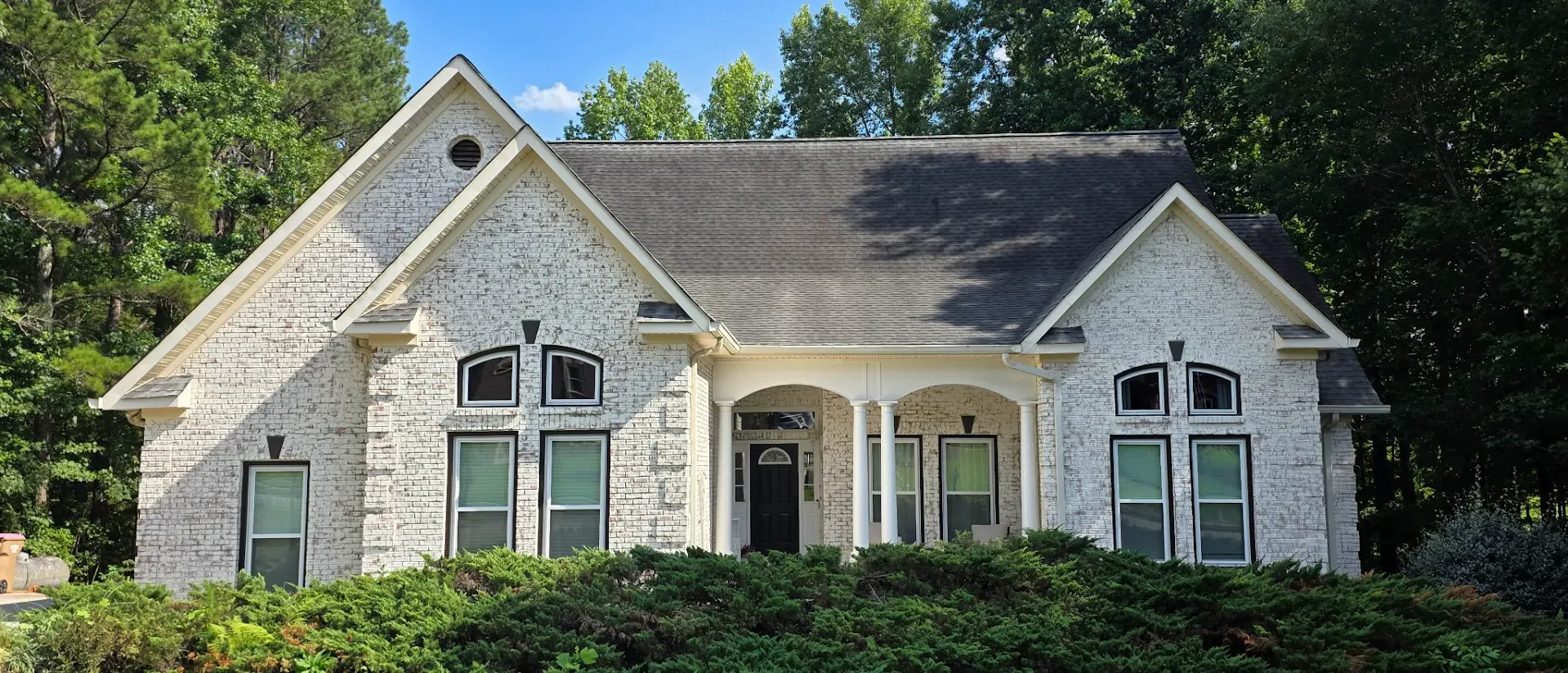 Stone house with green shutters and a front porch, surrounded by trees and shrubs