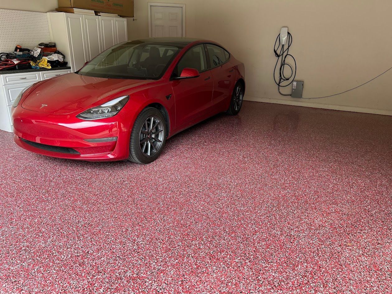 Red Tesla car parked in a garage with a red and white flecked floor next to a charging station.