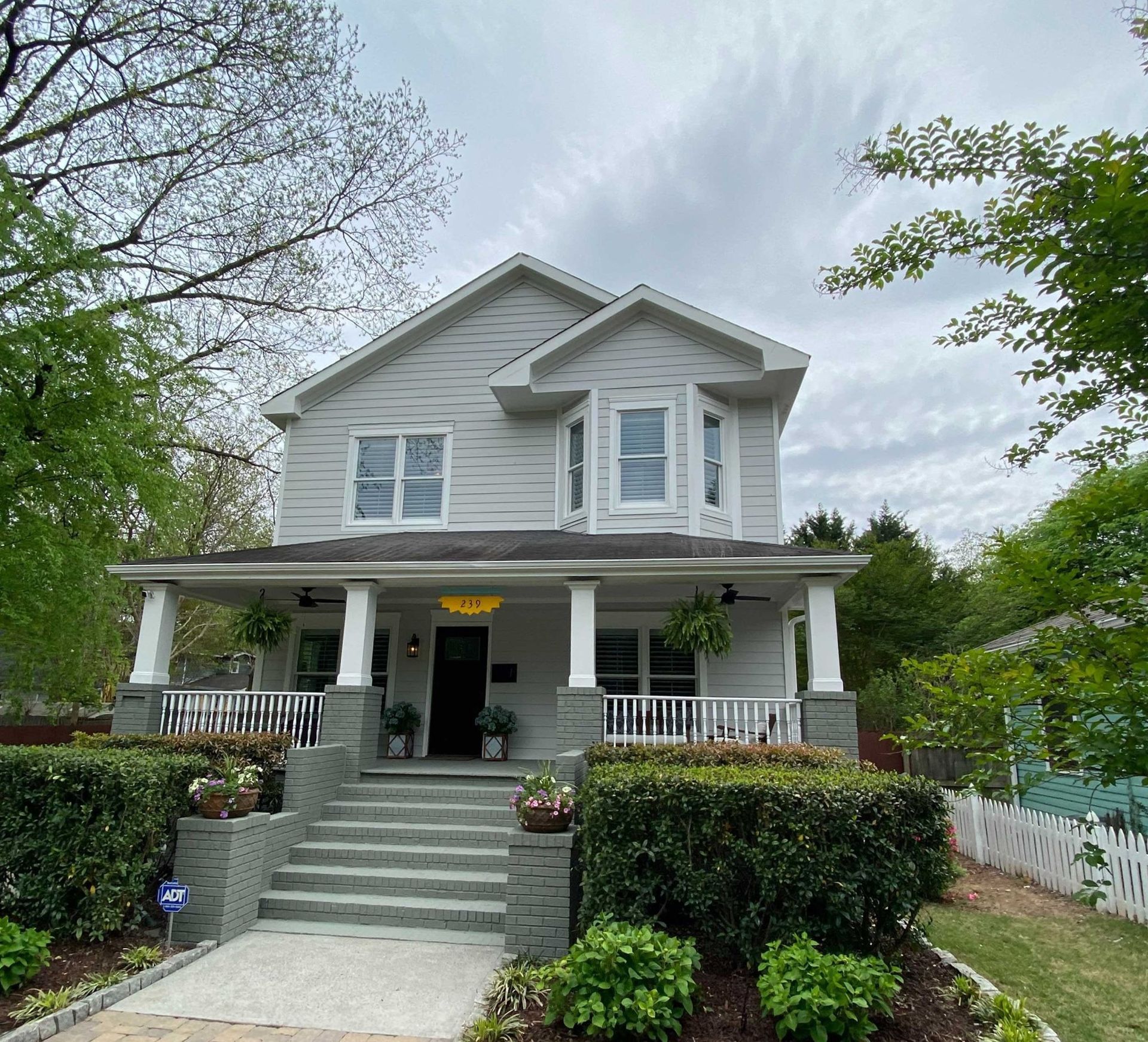 Two-story gray house with porch and bay windows, surrounded by landscaping and green foliage.