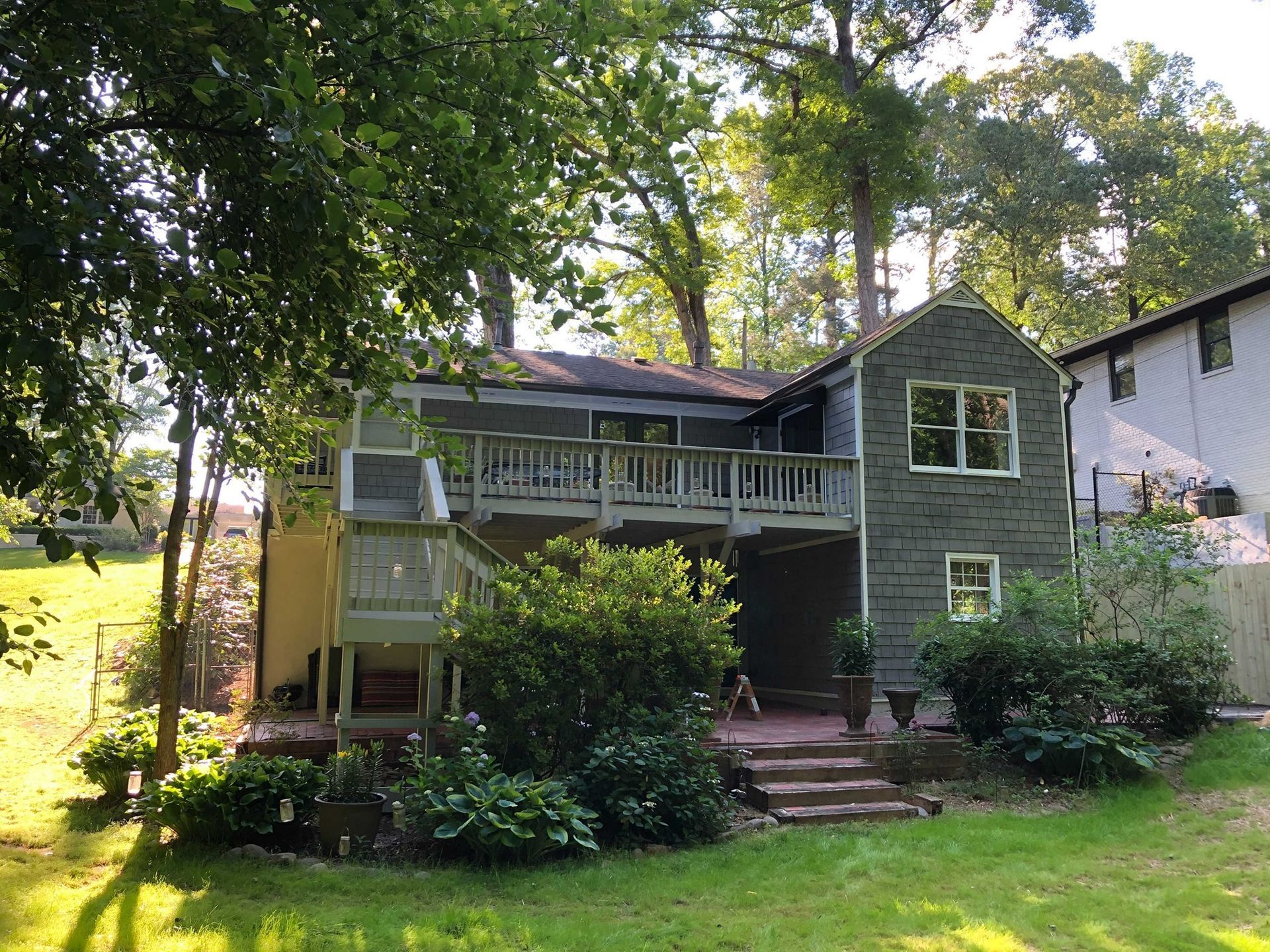 House with stone facade and large wooden deck, surrounded by lush greenery.