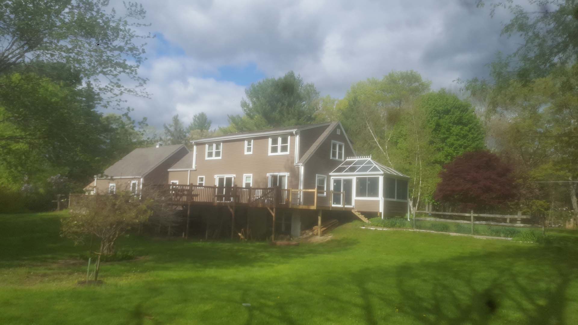 A large house with a large deck in the middle of a lush green field surrounded by trees.