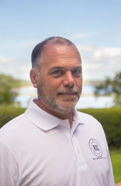 Man wearing a white polo shirt smiles in an outdoor setting with water in the background.