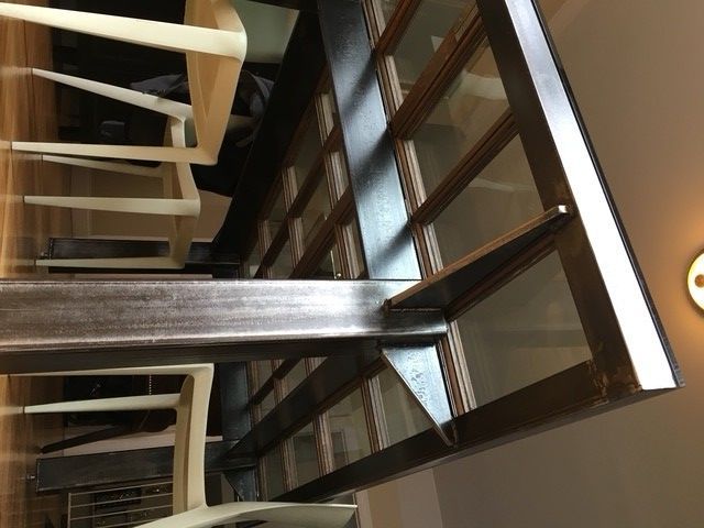A view from underneath a dark metal table frame with a glass top, featuring a central support beam and white chairs.