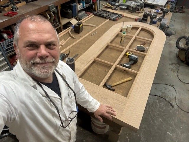 A smiling person in a white workshop coat standing by a newly crafted wooden arched door frame in a woodworking shop.