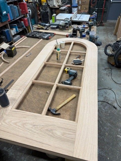 A wooden arched door frame with glass panel grid, under construction on a workbench in a woodworking shop.