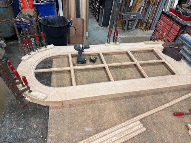 A wooden arched door frame being constructed in a workshop, clamped with tools and assembly parts on a workbench.