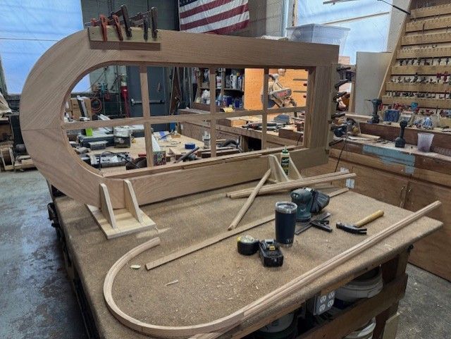 A large arched wooden window frame sits on a workbench in a workshop, with a matching curved trim piece in the foreground.