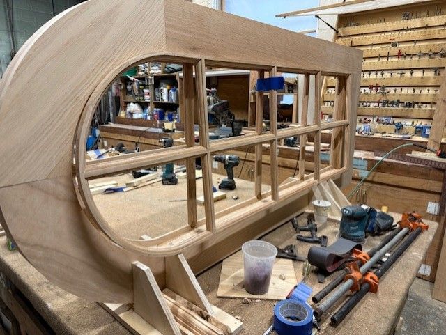 A partially constructed custom wooden window frame sits on a workshop workbench surrounded by various woodworking tools.