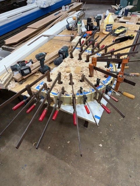 A curved wood piece clamped to a workbench in a workshop, surrounded by multiple tools and woodworking supplies.