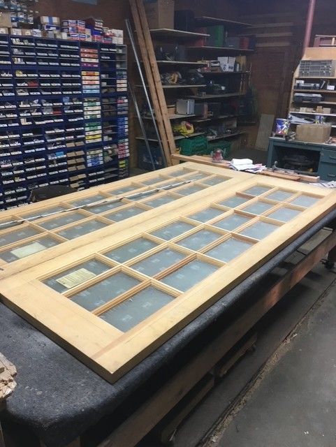 Two unfinished wooden French doors with glass panes lying flat on a workbench in a cluttered workshop.