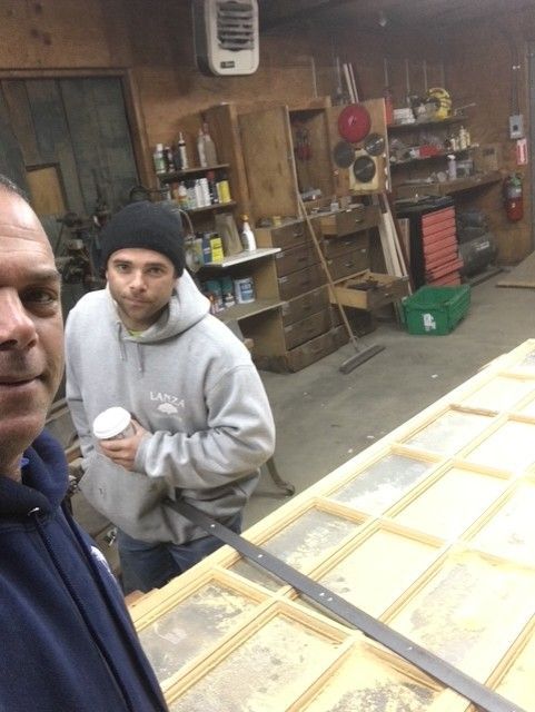 Two people in a workshop with a wooden grid frame, looking at the camera. One holds a coffee cup in a gray hoodie.
