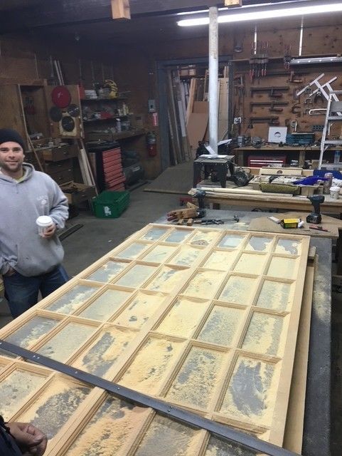 A person in a workshop stands next to a large wooden door frame laid flat on a workbench, surrounded by woodworking tools.