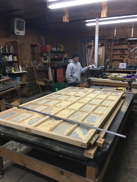 A person in a workshop standing behind a large wooden frame structure resting on a workbench.