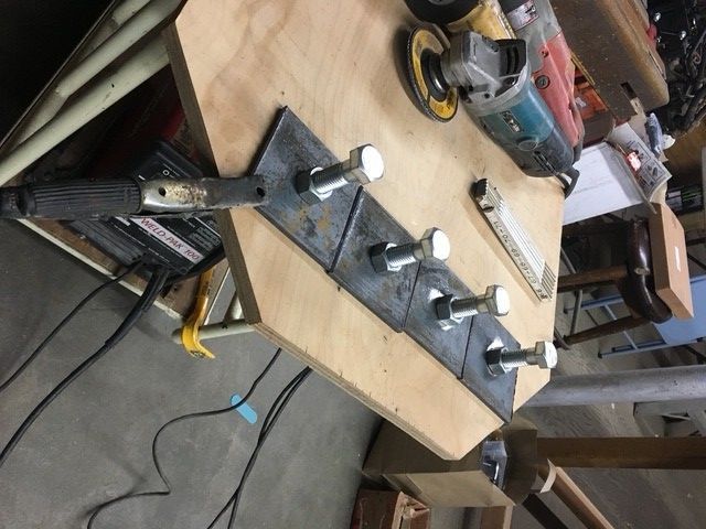 A welding ground clamp attached to a series of metal plates with bolts on a wooden workbench in a workshop.