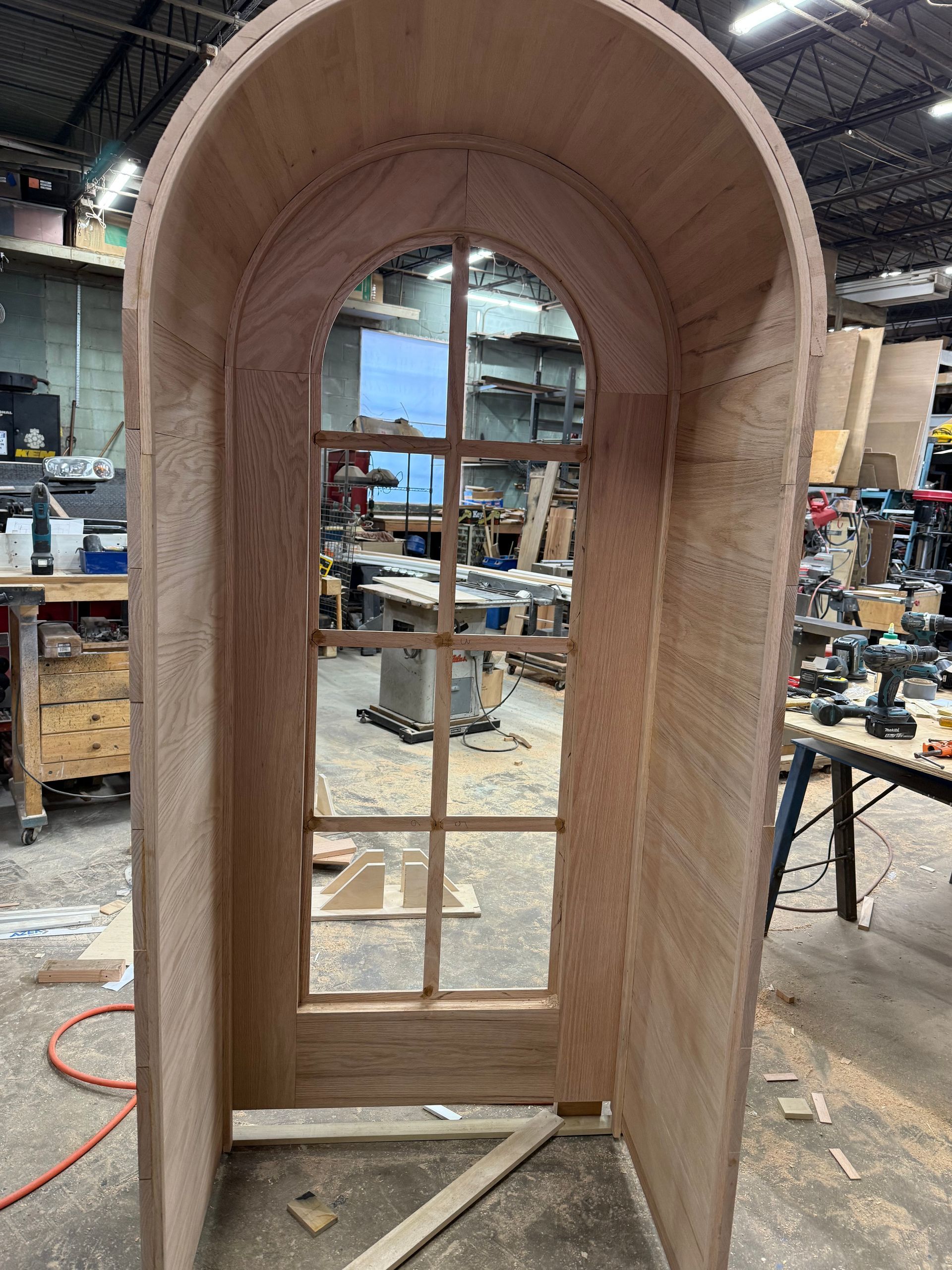 An unfinished, arched wooden door frame with a glass windowpane insert sitting in a woodworking workshop.