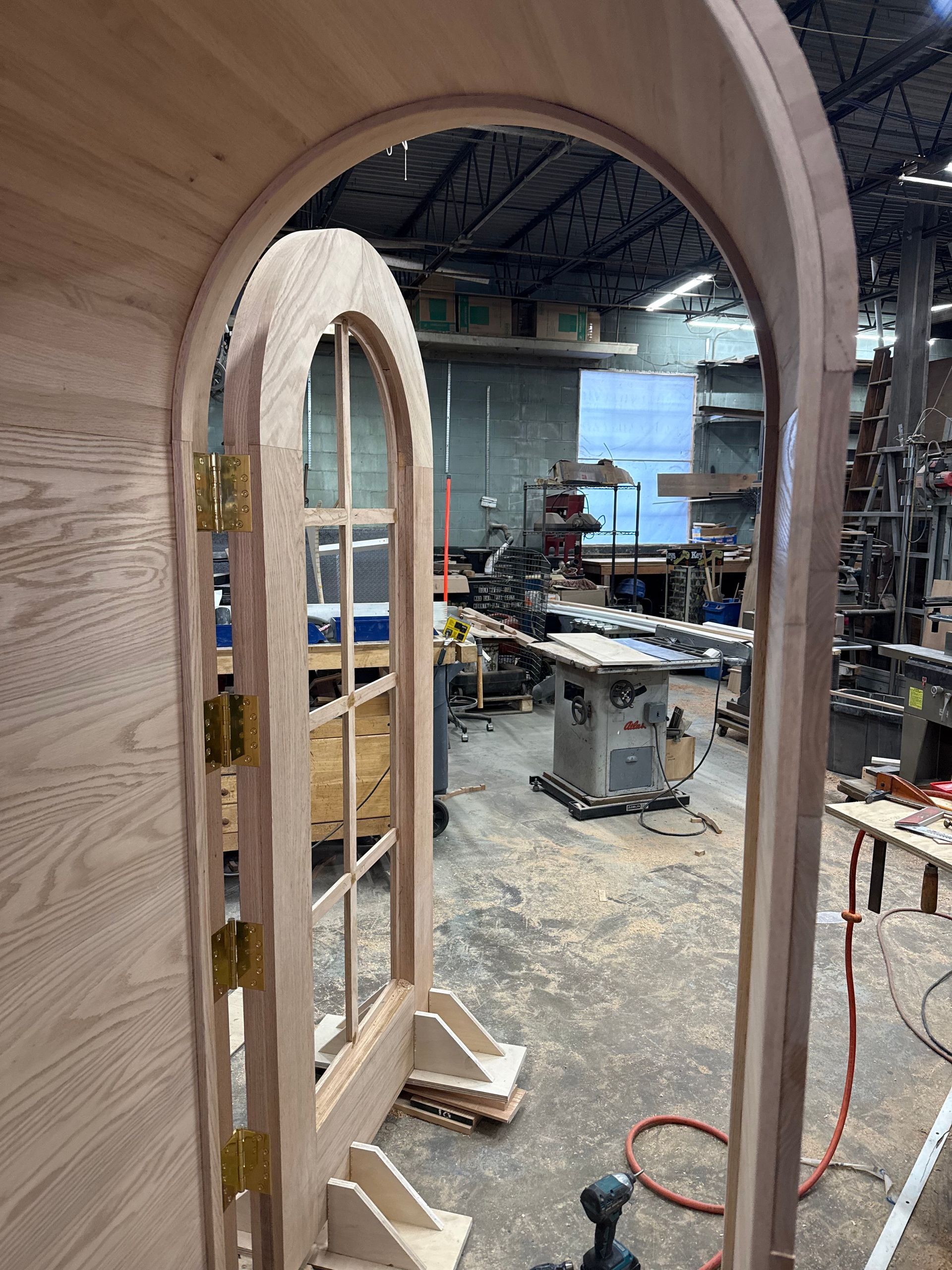 An arched wooden door frame stands in a woodshop, revealing a workshop area with tools and equipment in the background.