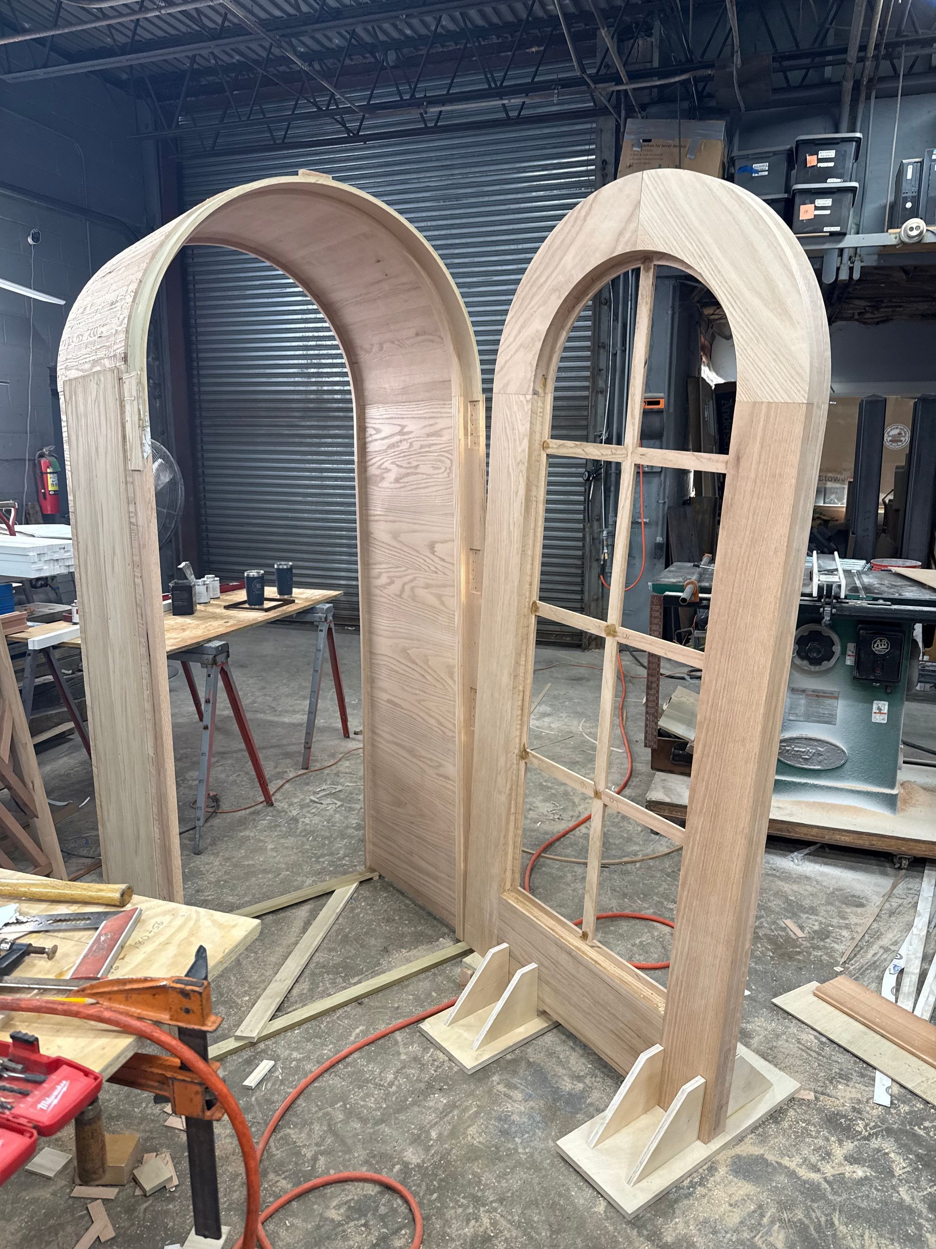 Two arched wooden frames stand in a woodworking shop, one with a grid window insert and one open-backed.
