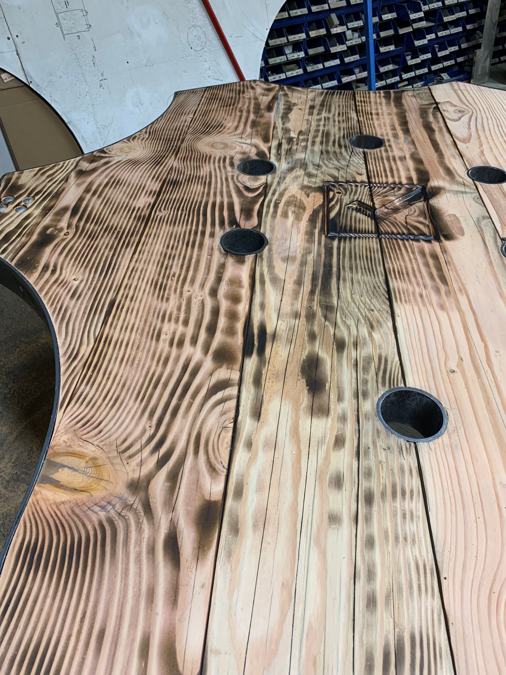 A close-up of a rustic, torched-wood surface featuring prominent grain patterns, dark burn marks, and several circular holes.