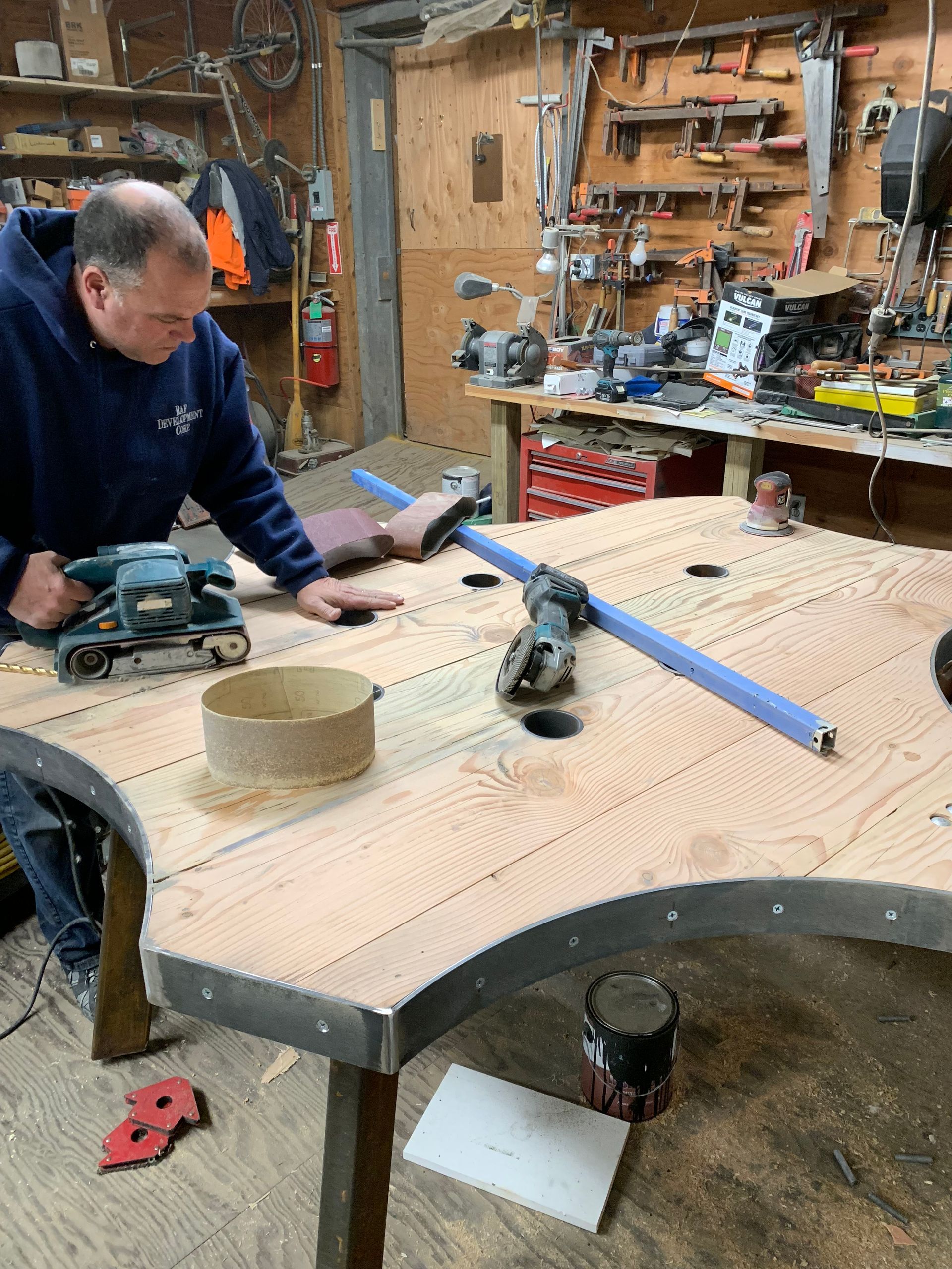 A person uses a belt sander on a large, curved wooden table surface in a cluttered workshop.