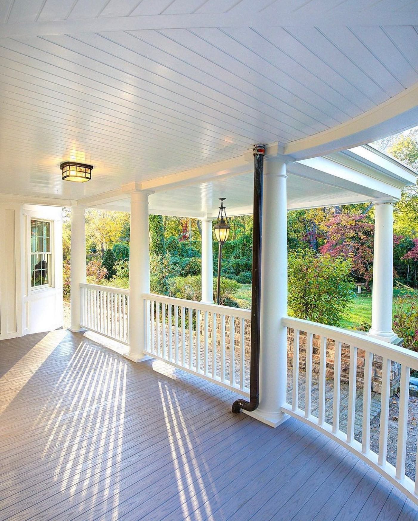 White porch with columns, railing, and a gray floor. Black rain gutter, light fixtures, and trees are visible in the background.