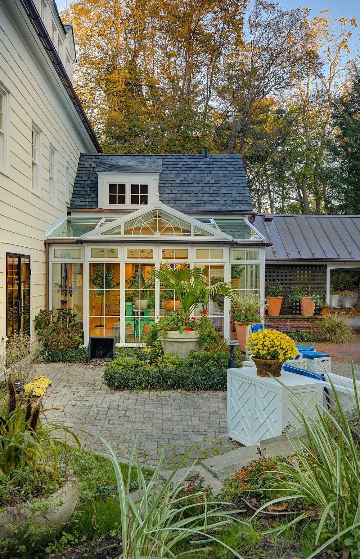 Sunroom with glass walls, attached to a white house. Plants and pots sit outside on a patio.