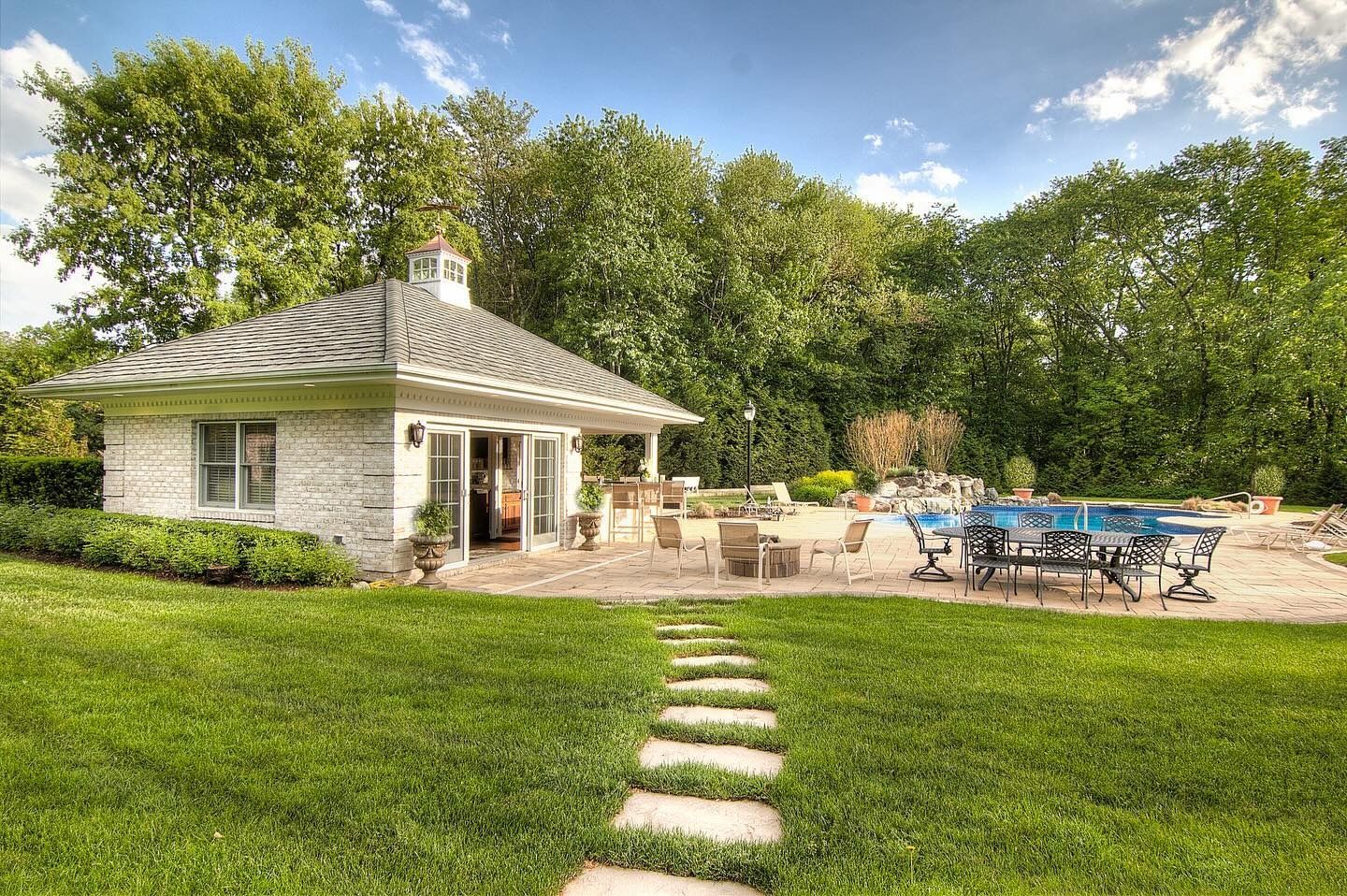 White cottage with open doors, patio with tables and chairs, green lawn, stone path.