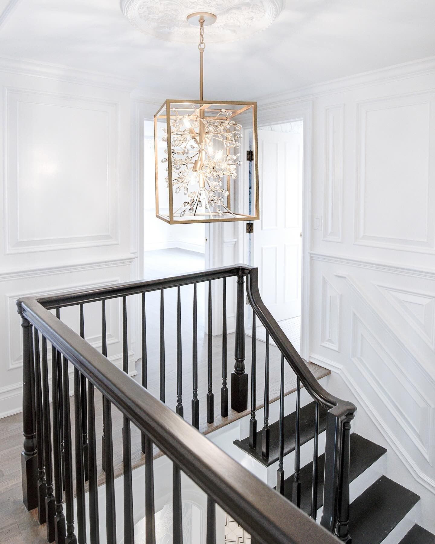 Staircase with black railing, white walls with molding, and a gold geometric chandelier.