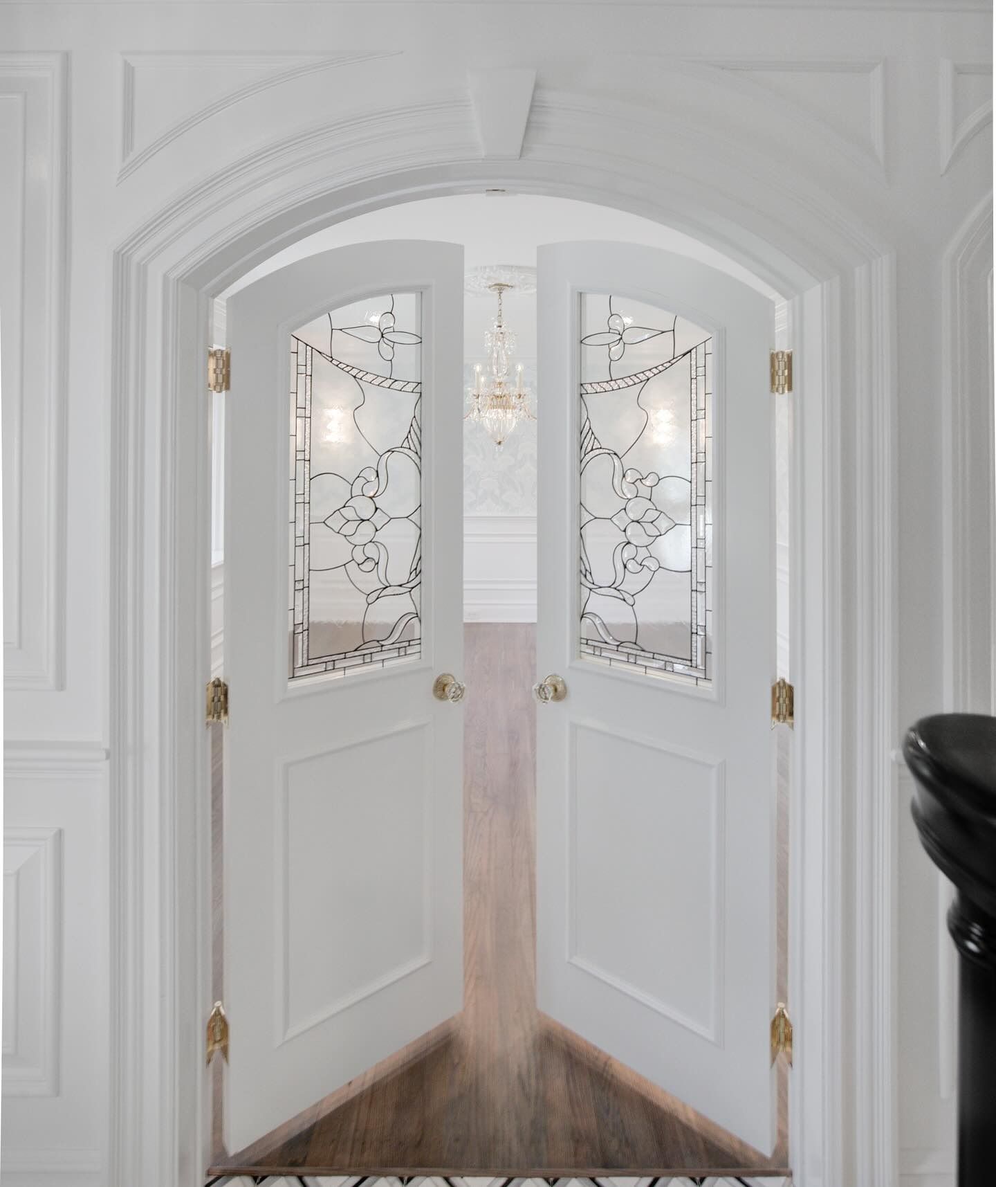 White, arched double doors with decorative glass panels, leading to a room with a chandelier.