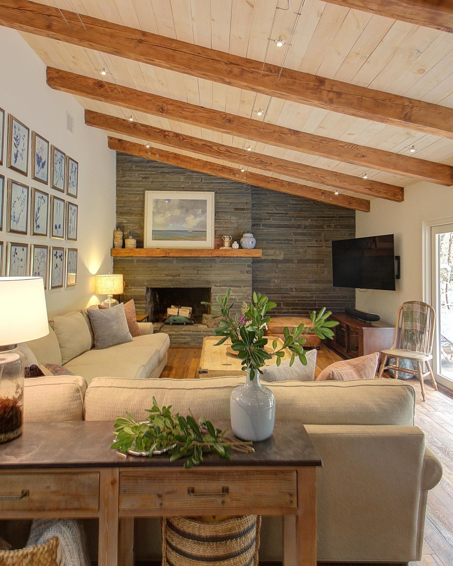 Living room with angled wood beam ceiling, stone fireplace, neutral-toned furniture, and large windows.